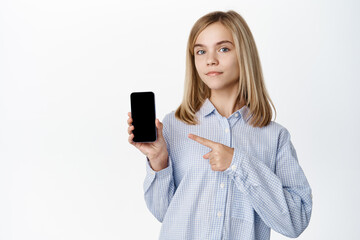 Cute blond girl, child pointing and showing on smartphone screen, empty mobile phone display, demonstrating app interface, white background