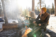Man Cooking Sausages Over Fire With Family In Snowy Forest