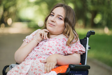 Portrait of beautiful woman with hairstyle looking at camera while sitting in wheelchair among green summer park. Lifestyles of people with chronic health disease.