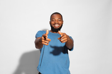 Black man with a blue t-shirt smiling and pointing fingers towards camera with a white background.