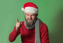A Man With A Beard In A Red Sweater And A Santa Claus Hat Looks Questioningly With A Cunning And Stern Look And Threatens With His Index Finger