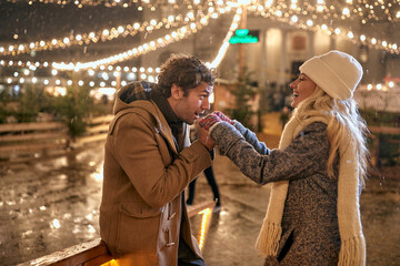 Wall Mural - A young guy is warming up the hands of his girl while they walking under christmas lights in the city. Christmas, New Year, holiday, love