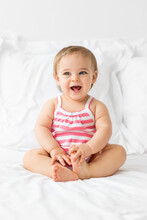 Smiling Baby Girl Sitting On A White Bed With Her Mouth Open
