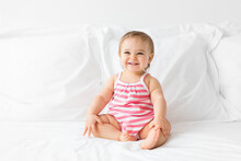 Happy Baby Girl Sitting On A White Bed Looking Away