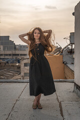 Wall Mural - Vertical shot of a young woman with a black dress and long hair posing on the roof of a building