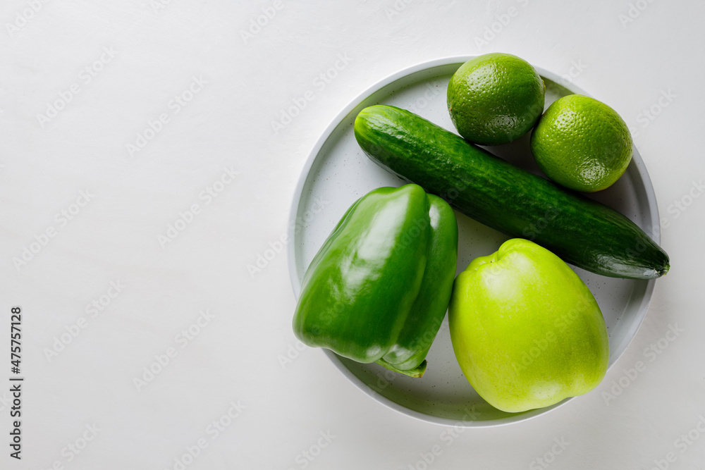 Various green vegetables and fruits on a white plate. Green apple, cucumber, bell pepper and lime on a white table. Copy space. Flat lay