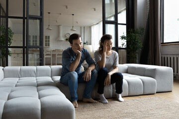 Fototapete - Tired unhappy young couple sitting on couch at home, thinking about relationship problems after quarrel, offended pensive girlfriend touching forehead, lost in thoughts, break up or divorce concept