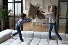 Overjoyed Little Sister And Brother Siblings Pillow Fighting, Jumping On Cozy Couch At Home, Happy Smiling Girl And Boy Having Fun, Engaged In Funny Activity, Spending Leisure Time Together