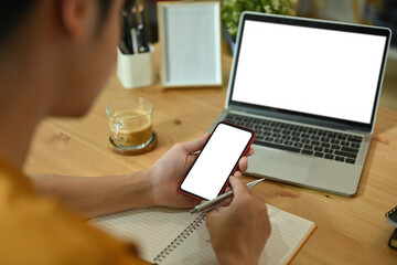 Poster - Close-up image of a freelance man holding a white blank screen smartphone and pen over the notebook on the wooden working desk surrounded with a white blank screen computer laptop and coffee cup.