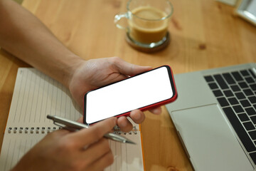 Poster - Close-up image of a freelance man holding a white blank screen smartphone and pen over the notebook on the wooden working desk surrounded with a white blank screen computer laptop and coffee cup.