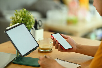 Poster - Photo of a young freelance man holding a white blank screen smartphone while taking notes at the wooden table surrounded by personal equipment and white blank screen digital tablet on tablet holder.