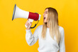 © luismolinero - Young blonde woman isolated on yellow background shouting through a megaphone to announce something in lateral position
