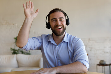 Happy sincere young Caucasian man in wireless headphones waving hand looking at camera, starting vide conference call conversation, communicating distantly or passing job interview, sitting at table.