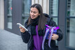 © puhimec - Young woman with a purple gift in hands uses a smartphone outside.