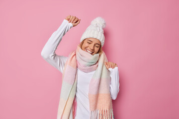 Wall Mural - Carefree optimistic woman wears white hat warm winter scarf around neck dances with joyful expression moves with rhythm of music isolated over pink background. Sincere emotions and feelings concept