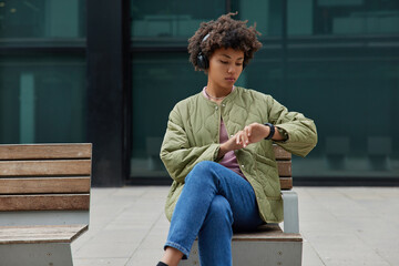 Wall Mural - Outdoor shot of serious woman with curly hair wears jacket and jeans sits on bench in urban area focused at smartwatch listens favorite music from playlist in headphones spends free time at city