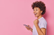 © WHstudio Leushin N - Happy joyful woman with curly hair holds mobile phone and celebrates win makes fist bump laughs has surprised expression wears casual t shirt stands sideways against pink background empty space
