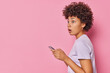 Sideways shot of stunned curly haired woman has shocked concerned expression wears t shirt accidentaly sent message via cellphone wrong person isolated over pink background woth copy space area