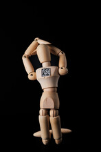A Wooden Mannequin With A QR Code Is Kneeling On A Dark Background. Selective Focus. The Concept Of The Trend Of Assigning QR Codes To People. Metaphor Is The Leash Of Global Digitalization.
