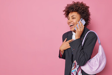 Wall Mural - Joyful optimistic curly haired woman in formal clothes giggles happily has telephone conversation carries bag holds smartphone near ear isolated over pink background with empty space for your text