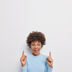 Wall Mural - Surprised cheerful woman with curly hair points above with both index fingers shows awesome advertisement wears transparent glasses and blue jumper cannot believe her eyes isolated on white wall
