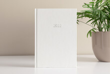 White Book Mockup, Diary For 2022 And Green Plant In Pot On White Table Against Background Of Beige Wall. Front View. Place For Text, Copy Space, Mockup