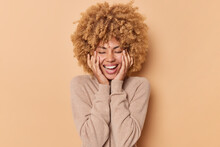 Happy Woman With Curly Hair Keeps Hands On Cheeks Closes Eyes And Smiles Joyfully Wears Casual Jumper Being In Good Mood Isolated Over Beige Background Recalls Nice Memories. Emotions Concept