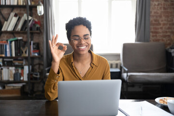 Happy young African American woman in glasses showing okey gesture, looking at computer screen, using sign lecture communicating by video conference call, sitting at table in modern loft style office.