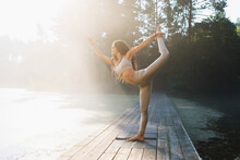 Woman Performs The Natarajasana Exercise, The Pose Of The King Of Dancers With An Outstretched Arm, Stands On A Wooden Bridge In A Park By A Pond On A Sunny Summer Morning, Trains In Beige Sportswear