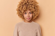 © WHstudio Leushin N - Portrait of serious confident sassy good looking woman with bushy curly hair looks directly at camera stands indoor against beige background wears casual jumper. Human face expressions concept