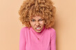 Photo of irritated young curly haired woman clenches teeth with hateful expression smirks face dressed in casual pink turtleneck isolated over beige background. Negative human emotions concept