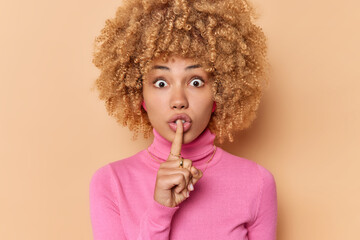 Wall Mural - Surprised curly haired woman presses index finger over lips asks to keep promise has shocked expression makes silence gesture dressed in casual pink turtleneck isolated over beige background