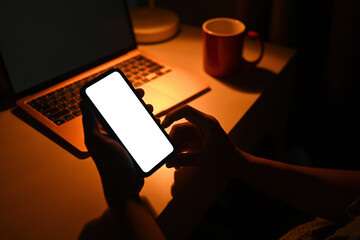 Poster - Photo of a hand using a white blank screen smartphone in the dark at the working desk surrounded by a computer laptop and coffee cup.