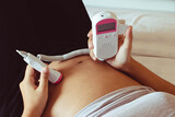 Fototapeta Zachód słońca - The close up pregnant woman using pocket fetal doppler to monitor baby heart beat.