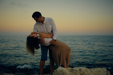 Man Tilts Woman Backward Holding Her On Arms Opposite Seaside Sunset. Young Couple On A Date. Romantic Seaside Date