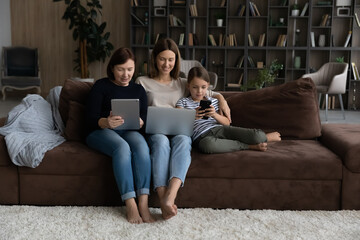 Wall Mural - Family online. Three women relatives of diverse age sit on sofa at living room use digital gadgets. Old grandmother young adult mother preteen schoolgirl daughter grandchild hold laptop touchpad phone