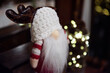 Ozdoba świąteczna, skrzat w ciepłej czapce, z białą brodą i porożem renifera. W tle światełka choinkowe