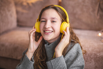 Wall Mural - Cheerful young woman listening to music in headphones sitting near the sofa in the room