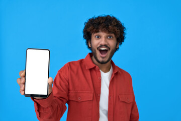 Excited young indian man holding cellphone showing big white blank empty mockup screen. Surprised guy advertising betting app on phone isolated on blue background. Smartphone application ads