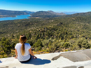 Wall Mural - Female sitting on the concrete ground looking at the dense forest and blue lake under a clear sky