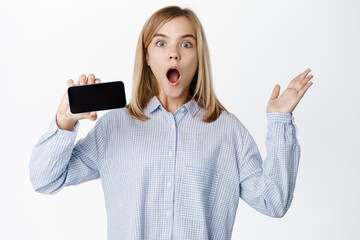 Enthusiastic blond little girl, showing horizontal smartphone screen, empty mobile phone display, app interface, standing over white background