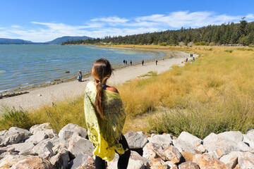 Poster - Brunette female standing on the stones looking at the beach with people under a blue sky