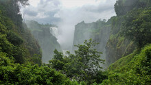 Victoria Falls Streams Collapse Into A Narrow Gorge With Steep Rocky Slopes. Thick Fog Over The Abyss. Cloudy. In The Foreground Is Lush Green Vegetation. Zimbabwe