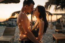 Love On The Beach. Loving Couple In The Evening With Lights On The Neck Hugging Tropical Resort. Guy With Girl Celebrate Christmas Or Valentine's Day