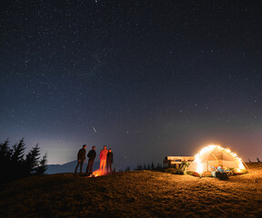 Wall Mural - Group of friends standing at night under starry sky on green mountain lawn and looking at flame of campfire in camp, next to shining brightly tent and wooden table. Night camping on mountain hills.