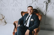 © shchus - Stylish groom sit on a leather armchair in a modern studio against a background of a white wall. Wedding portrait.