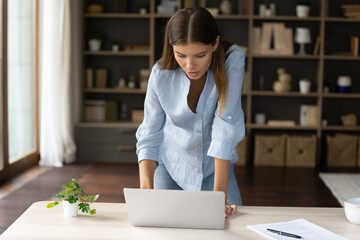 Wall Mural - Serious young employee girl finishing work task in hurry, standing and using laptop in home office, leaning on desk, typing, looking at screen, talking on video call, using online virtual app.