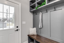 A Staged, Gray Mudroom / Entryway With Bench Seating, Coat Hooks, And Storage Above. A Scarf Hangs From A Hook And A Plant Sitting On A Shelf.