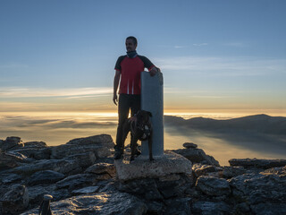 Caucasian male standing next to a dog and a rocky pole on the top of at sunset with a cloudy sk