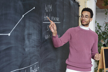 Poster - Young confident teacher of economics pointing at graph on blackboard while explaining it to colleagues at seminar